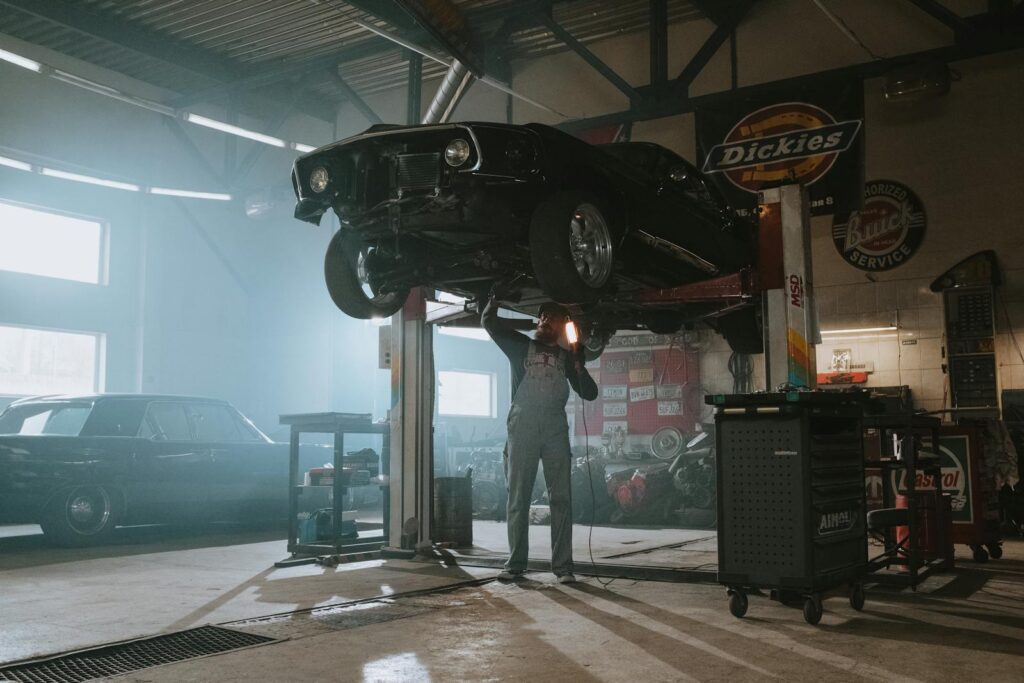 Male mechanic using a light to inspect a classic car lifted in an automotive garage.